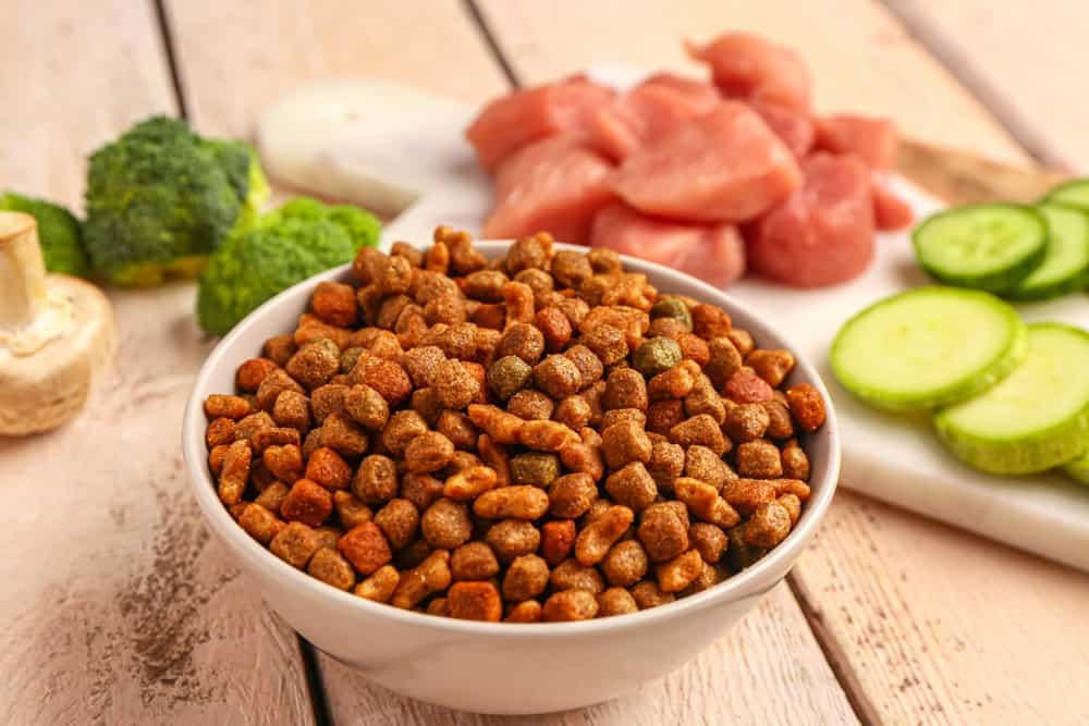 A bowl of brown and orange kibble sits on a wooden table. Behind it, there is a cutting board with chunks of raw chicken, sliced cucumbers, broccoli, and a mushroom. The scene suggests ingredients for preparing a pet's meal.